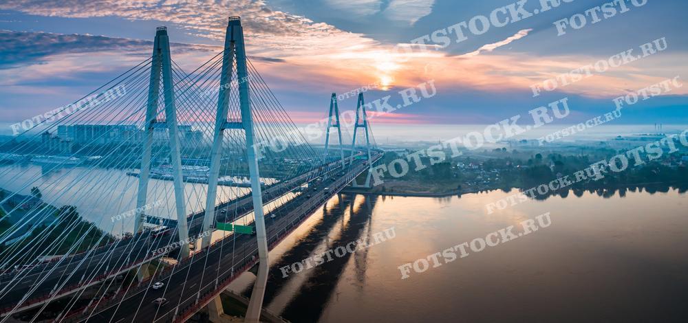 На фотографии изображен Вантовый мост (Большой Обуховский мост) в Санкт-Петербурге, пересекающий реку Неву. Утренний туман создает таинственную атмосферу, а восходящее солнце придает небу розовые и оранжевые оттенки. С воды поднимается легкая дымка, отражающая свет солнца и создающая живописный вид.